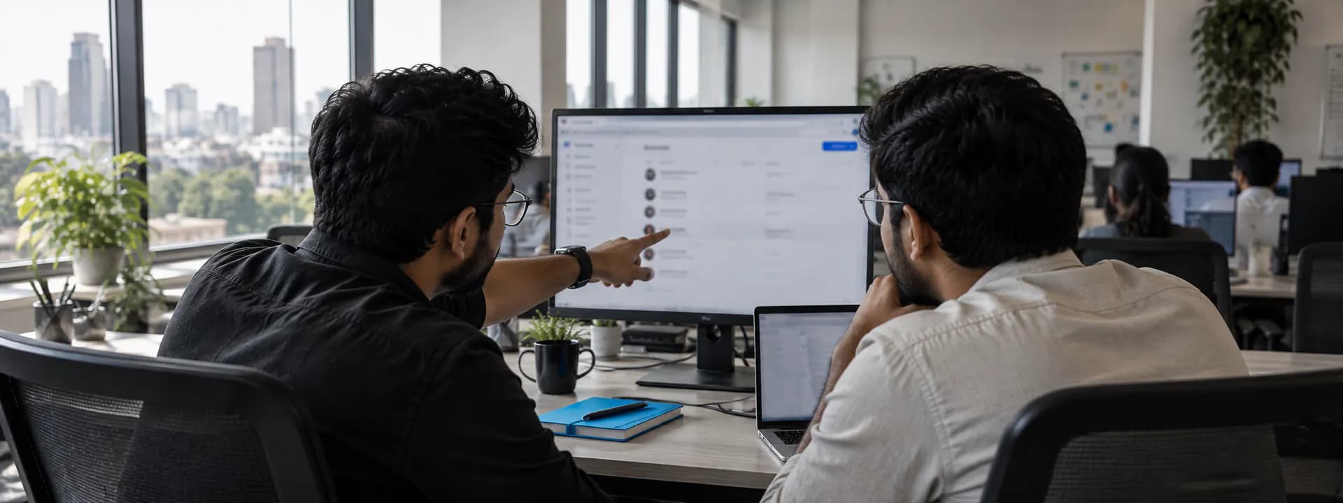 Two engineers at a desk in the QalbIT office, one pointing at a monitor showing a repository settings page with the client organisation as owner.